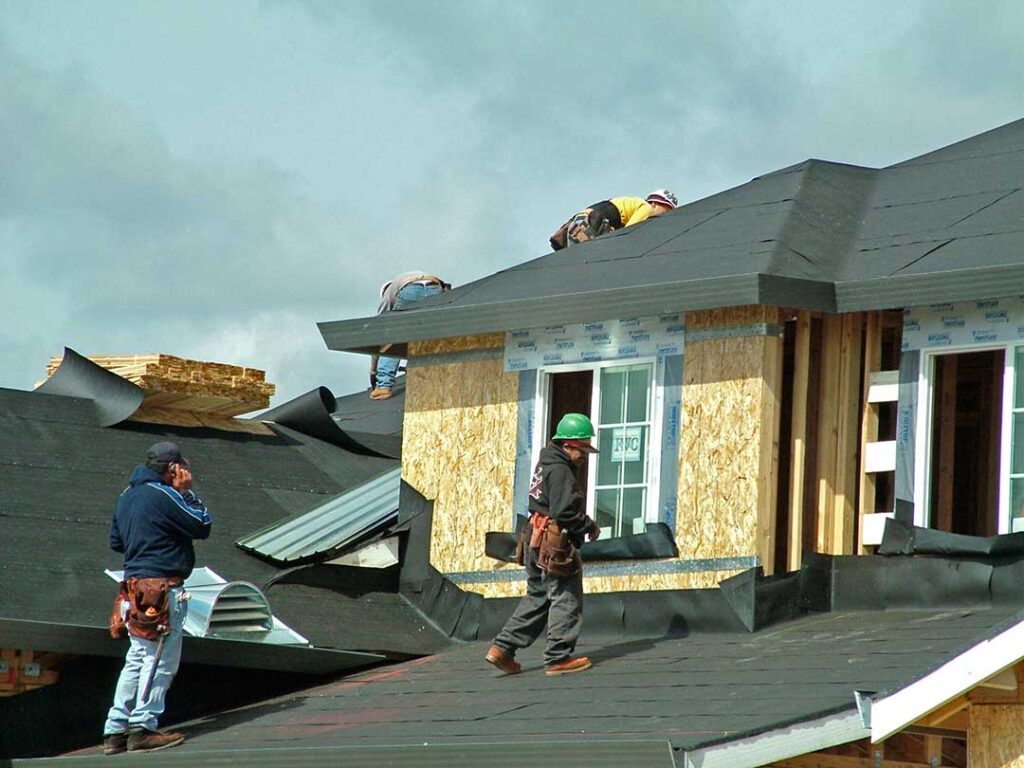 A worker installs attic insulation beneath roof rafters on a sunny day, with tools and materials visible.