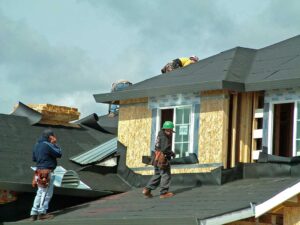 A worker installs attic insulation beneath roof rafters on a sunny day, with tools and materials visible.